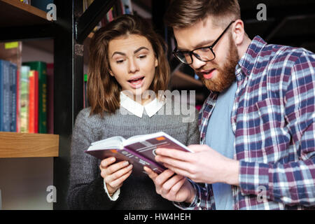 Man and woman students standing by bookshelf in library and reading book Stock Photo