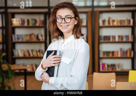 Portrait of a smiling young female student in eyeglasses holding book and looking at camera in library Stock Photo