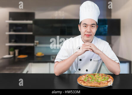 Asian young man chief in white uniform offers to you delicious pizza. Man chief preparing pizza in kitchen. Stock Photo