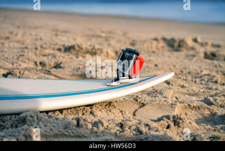 Surfboard with attached action camera lies on the sand on the beach. Action camera is in waterproof case for swimming. Stock Photo
