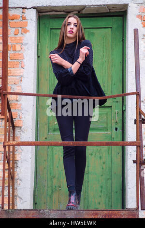 Full length of beautiful and slim girl with long hair posing against abandoned house and old green door. Stylish and young female in black posing with Stock Photo