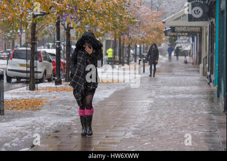 Cold & snowing, lady (people) wearing welly boots, scarf & winter coat (hood up) walks past shops in snow - The Grove, Ilkley, West Yorkshire, GB. Stock Photo