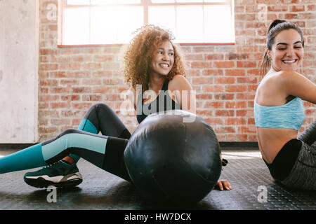 Two happy young women sitting on gym floor with medicine ball. Female friends taking a break from workout. Stock Photo