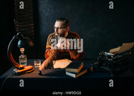 Thoughtful bearded writer in glasses sitting at the table. Retro typewriter, feather, crystal decanter, books and vintage lamp on the desk Stock Photo