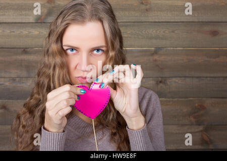 Young female with funny pink heart on a stick, wooden background. Fun photo props and accessories for shoots Stock Photo