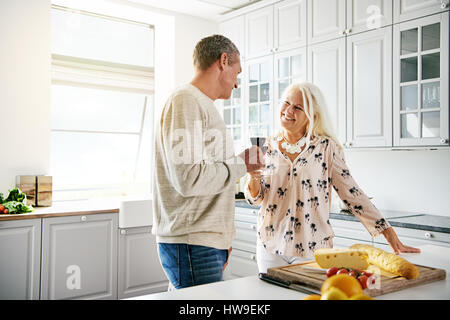 Loving retired couple preparing a meal together in the kitchen enjoying an aperitif drinking red wine while smiling and chatting Stock Photo
