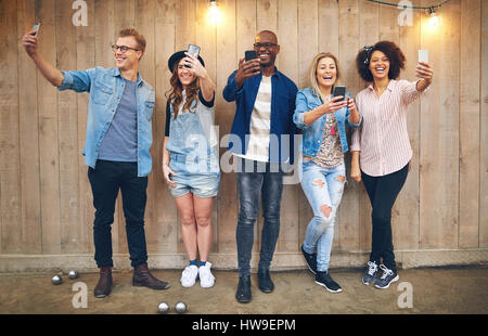 Group of happy young people standing against wooden unpainted wall with decorations, all taking selfies with their smartphones, smiling Stock Photo