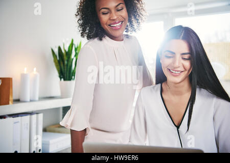 Happy successful young women entrepreneurs working together at a laptop computer in a high key office with sun flare, close up cropped view Stock Photo