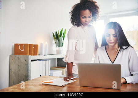 Two young female business partners working together using a laptop computer at the office with bright sun flare over their shoulders Stock Photo