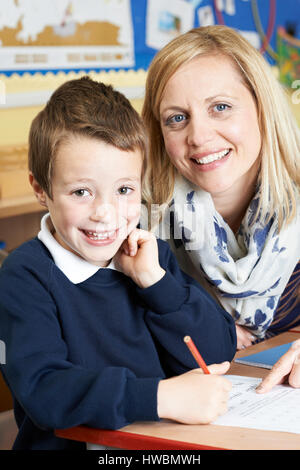 Teacher Helping Male Elementary Pupil Working At Desk Stock Photo
