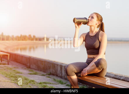 young girl of Caucasian appearance drinks a protein cocktail from a shaker after jogging in the open air. The bright sun illuminates it. Stock Photo