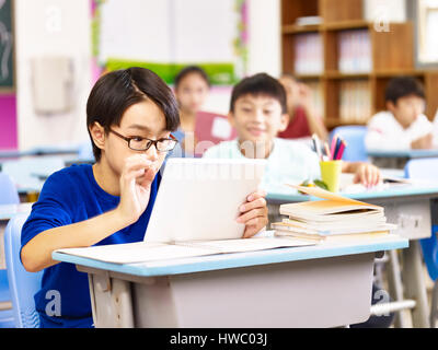 asian elementary schoolboy looking with curiosity at a tablet computer held by his classmate, focus on the boy in foreground. Stock Photo