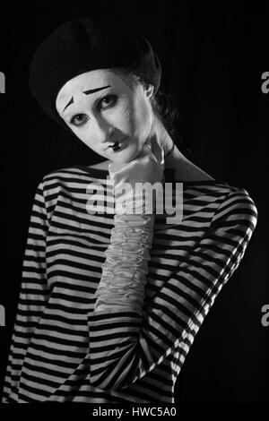 Emotional professional mime in stripped blacn and white shirt with black cap and white bow tie standing against black background in circus Stock Photo