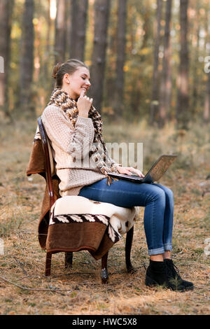 Interesting,attractive,cute girl with bob in the hair,in warm sweater and scarf sit on chair in the autumn forest outdoors.Girl is online,is on the In Stock Photo
