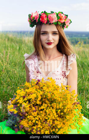 Beautiful fashionable attractive girl with big wreath of beautiful bright flowers on her hair, head. Sitting in field cute girl with big bouquet ful o Stock Photo