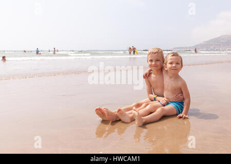 Two cute adorable little brothers boys sitting on the beach ocean sea