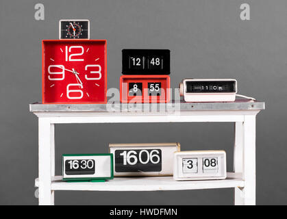 Still Life of Collection of Clocks in Variety of Styles - Clock Radio, Flip Style, and Alarm Clocks - on White Table in front of Gray Background Stock Photo