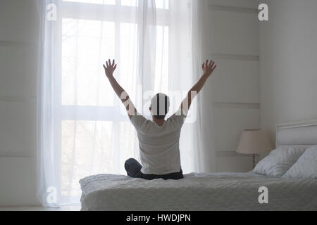 Happy young man stretching in bed after waking up, back view, total white concept Stock Photo