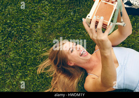 Young woman lying on grass taking selfie with retro camera Stock Photo