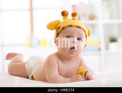 Funny baby boy in giraffe hat lying on his belly in nursery. Little kid lies on bed with opened mouth and holds soother in hands Stock Photo