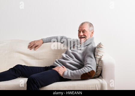 Senior man in gray sweater sitting on sofa, studio shot. Stock Photo