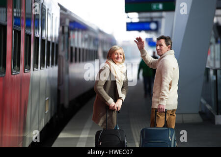 Senior couple on train station pulling trolley luggage, waving. Stock Photo