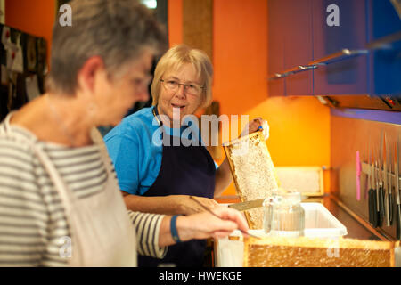 Two senior female beekeepers scraping honeycombs in kitchen Stock Photo
