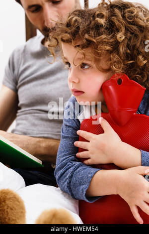 Girl hugging hot water bottle while father reads storybook in bed
