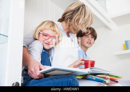 Mature woman sitting on kitchen counter reading storybooks with son and daughter Stock Photo