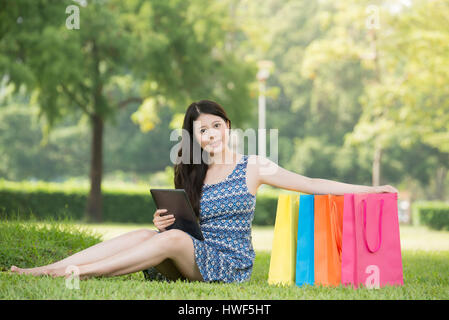 happy asian woman using digital tablet shopping online sitting on grass. outdoors background. technology and people concept Stock Photo