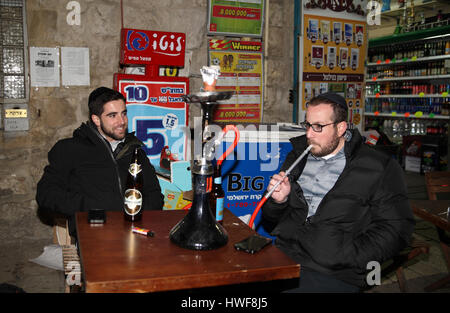 Two young American Jewish friends, tourists, sit at an outdoors Jerusalem pub or bar, one of them smokes a water pipe, the other one smiles at him. Stock Photo