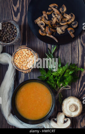 Pea soup in black plate. Fried mushrooms. Dried pea, pepper in glass jars.  Fresh parsley and rosemary. Dark wooden table. Toned. Stock Photo