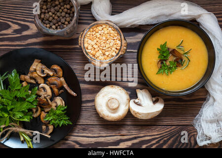 Pea soup with mushrooms in black plate. Fried mushrooms. Dried pea, pepper in glass jars.  Fresh parsley and rosemary. Dark wooden table. Toned. Stock Photo