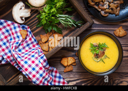 Pea soup with mushrooms in black plate. Fried mushrooms. Dried pea, pepper in glass jars.  Fresh parsley and rosemary. Rye crackers. Dark wooden table Stock Photo