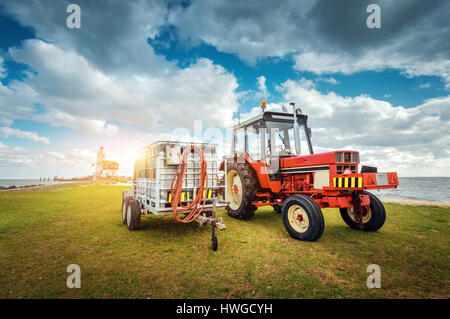 Red tractor with trailer on the grass field against lighthouse and blue cloudy sky in spring at sunset. Agricultural tractor. Agricultural machinery a Stock Photo
