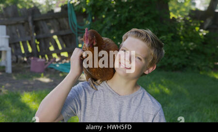 High angle portrait of smiling boy petting rooster on shoulder in backyard