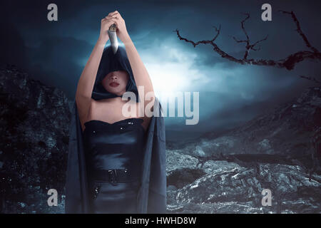 Witch woman with black costume wearing cloak and holding knife on the rock hills at the midnight Stock Photo