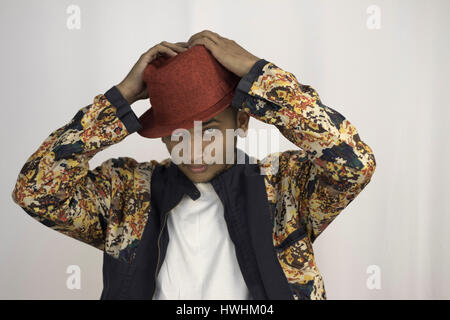 A young man with red hat and pixel printed multi colored jacket, posing with hands on the hat, making fashion statement Stock Photo