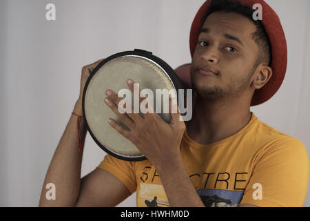 A young man with red hat, orange t-shirt, holding djembe on his right shoulder and playing it with left hand. (drumming) Stock Photo