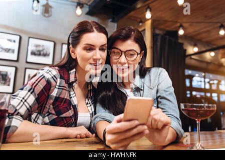 Selfie time. Positive delighted women wearing casual clothes keeping smile on faces while looking at mobile phone Stock Photo