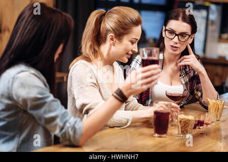 After workday. Delighted blonde sitting in semi position between two friends putting arms on the table while listening to her colleague Stock Photo