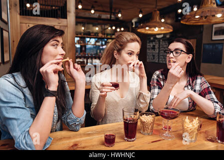 Tasty dish. Frustrated blonde covering her lips, sitting between her friends while holding glass with alcohol Stock Photo