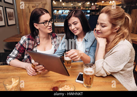 How do you do. Friendly girl wearing jeans jacket holding her tablet in right hand while sitting between her friends Stock Photo