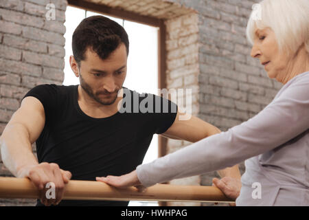 Working hard for success. Serious thoughtful concentrated dancer teaching elderly woman while standing next to the barre and demonstrating right postu Stock Photo