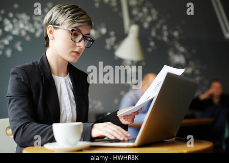 Profile view of confident young journalist with stylish haircut sitting in cafe with cup of coffee and writing article for magazine Stock Photo