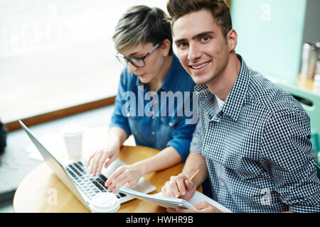 Handsome young manager looking at camera while his female colleague writing response email to their business partner Stock Photo