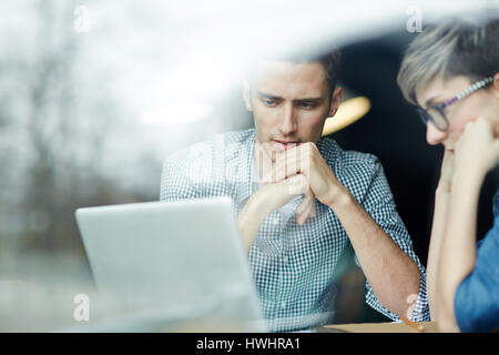 Concentrated university students sitting at table and watching video lecture on laptop, view through window Stock Photo