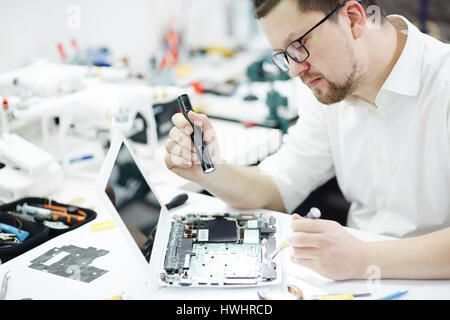 Side view portrait of man opening disassembled computer and using flashlight to inspect broken parts in circuit board at table in workshop Stock Photo