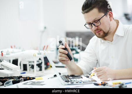 Side view portrait of man inspecting disassembled laptop with flashlight, looking for broken pats in workshop Stock Photo