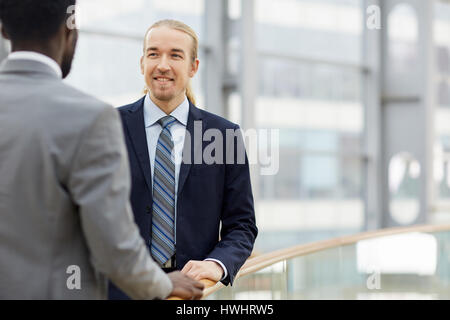 Portrait of two contemporary businessmen, one of them African, talking casually and smiling by glass railing in modern office building Stock Photo